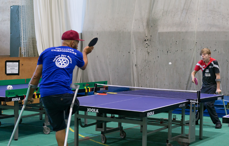 Two male-presenting disabled people are playing table tennis. Both are playing in standing positions assisted by crutches. The player nearest to the camera is a single-leg amputee. They have played the ball, and the other player is preparing to return it.