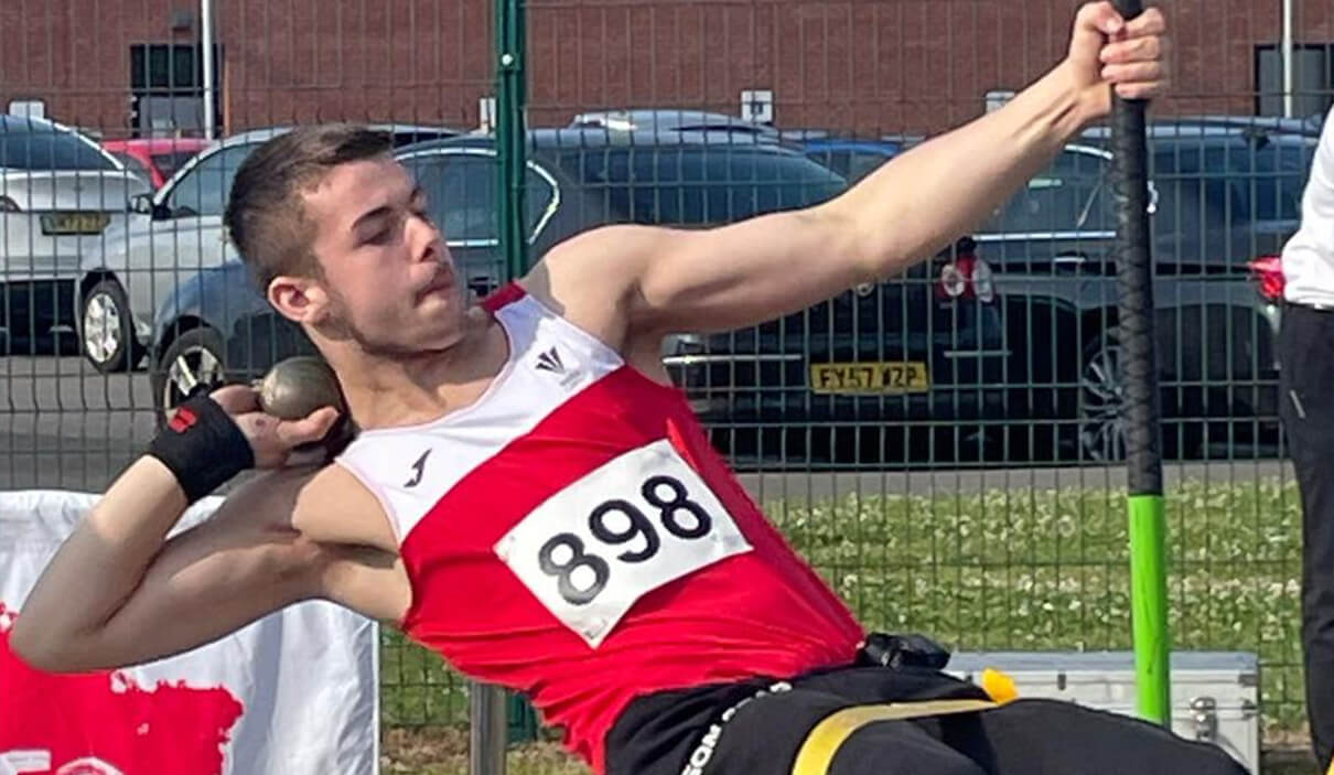 Jac is winding up for a seated shot put throw. He is in Welsh Athletics kit, with a competition number on his chest. He is holding on to a vertical bar to give him leverage to throw.