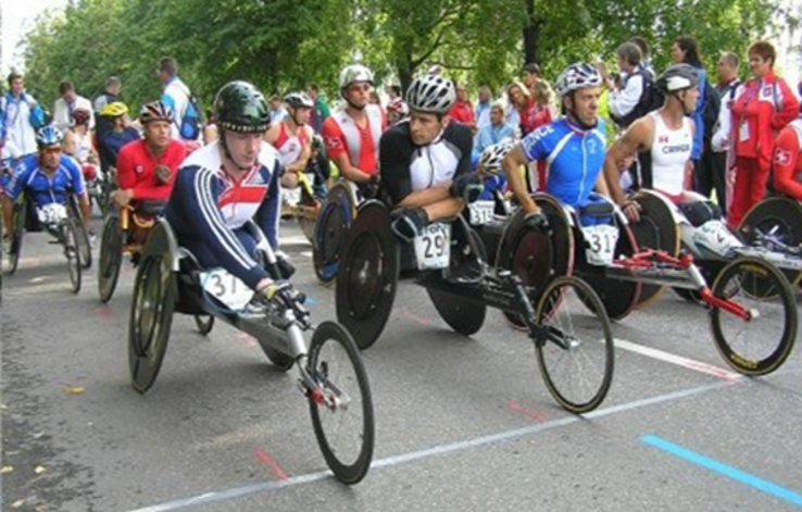 Professional para athletes line up to start a wheelchair race. They are wearing helmets, race numbers, and national-level kit (GB, Czech, France and Swiss kits are clearly visible). The race is outdoor, on a road, and there is a crowd of supporters.
