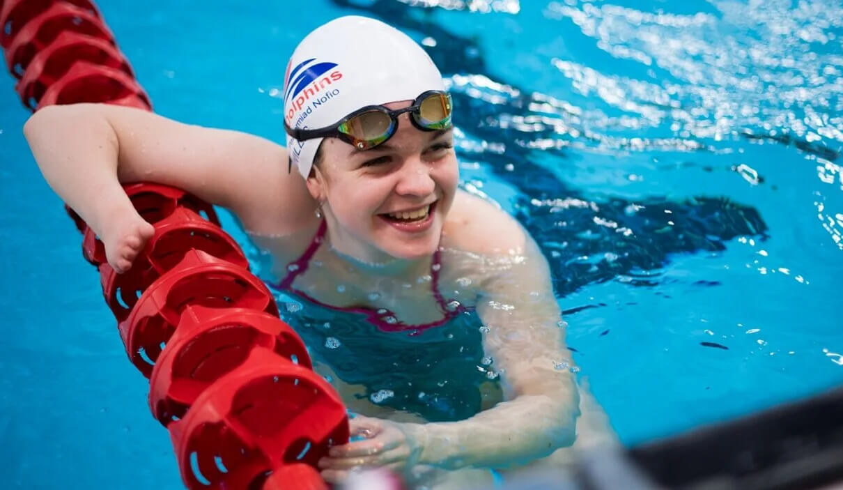 Meghan in a swimming pool. She is leaning on the lane dividers, smiling up and people on the side of the pool. She had lifted her goggle up, and is wearing a Dolphins cap.