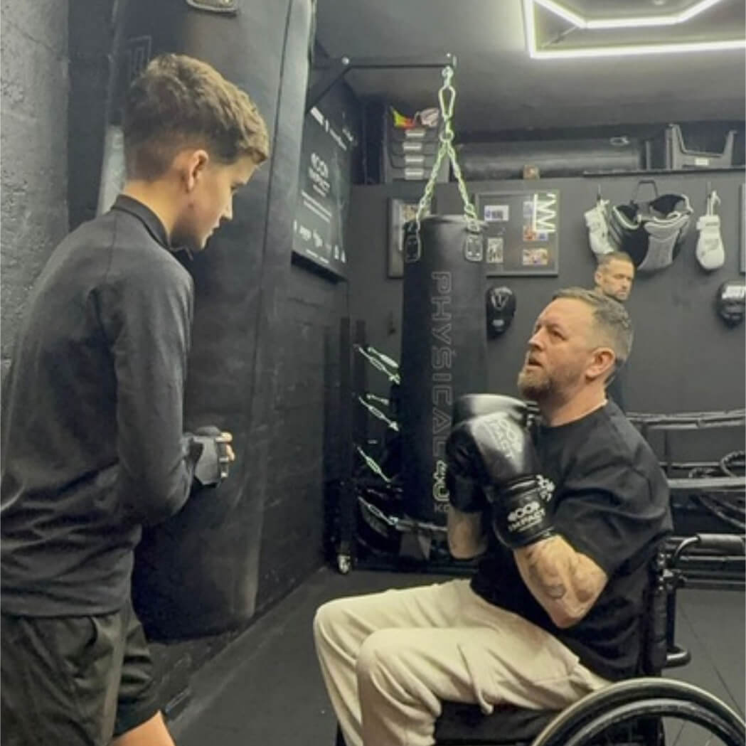 A chair user does boxing drills with a punching bag. They are supported by a coach who is holding the punching bag.