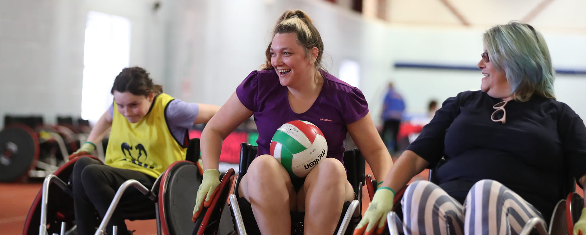 Three female-presenting participants play wheelchair rugby at insport Series: Para Sport Festival. The central participant has the ball in their lap, and gloved hands on both wheels of her sports wheelchair. She is smiling broadly, possibly laughing.