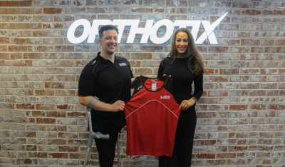 Nathan Stephens (Performance Pathway Senior Officer) & Hayley Huntley (Managing Director at Orthotix) stand in front of an illuminated Orthotix logo at Orthotix' headquarters in Cardiff, together holding up a Disability Sport Wales jersey.