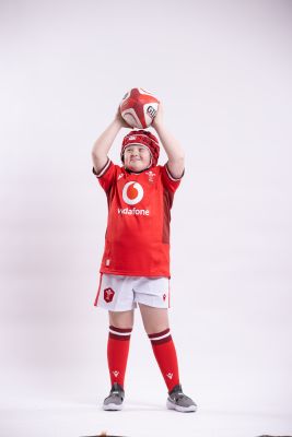 A young person wearing a full Wales Rugby kit holding a ball in the air and smiling