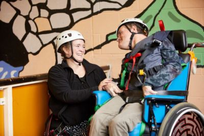 Photo from SENSE. A chair-using participant and non-disabled session facilitator smile at each other. They are wearing helmets and safety harness to support climbing and/or abseiling.
