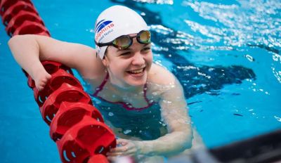 Meghan in a swimming pool. She is leaning on the lane dividers, smiling up and people on the side of the pool. She had lifted her goggle up, and is wearing a Dolphins cap.