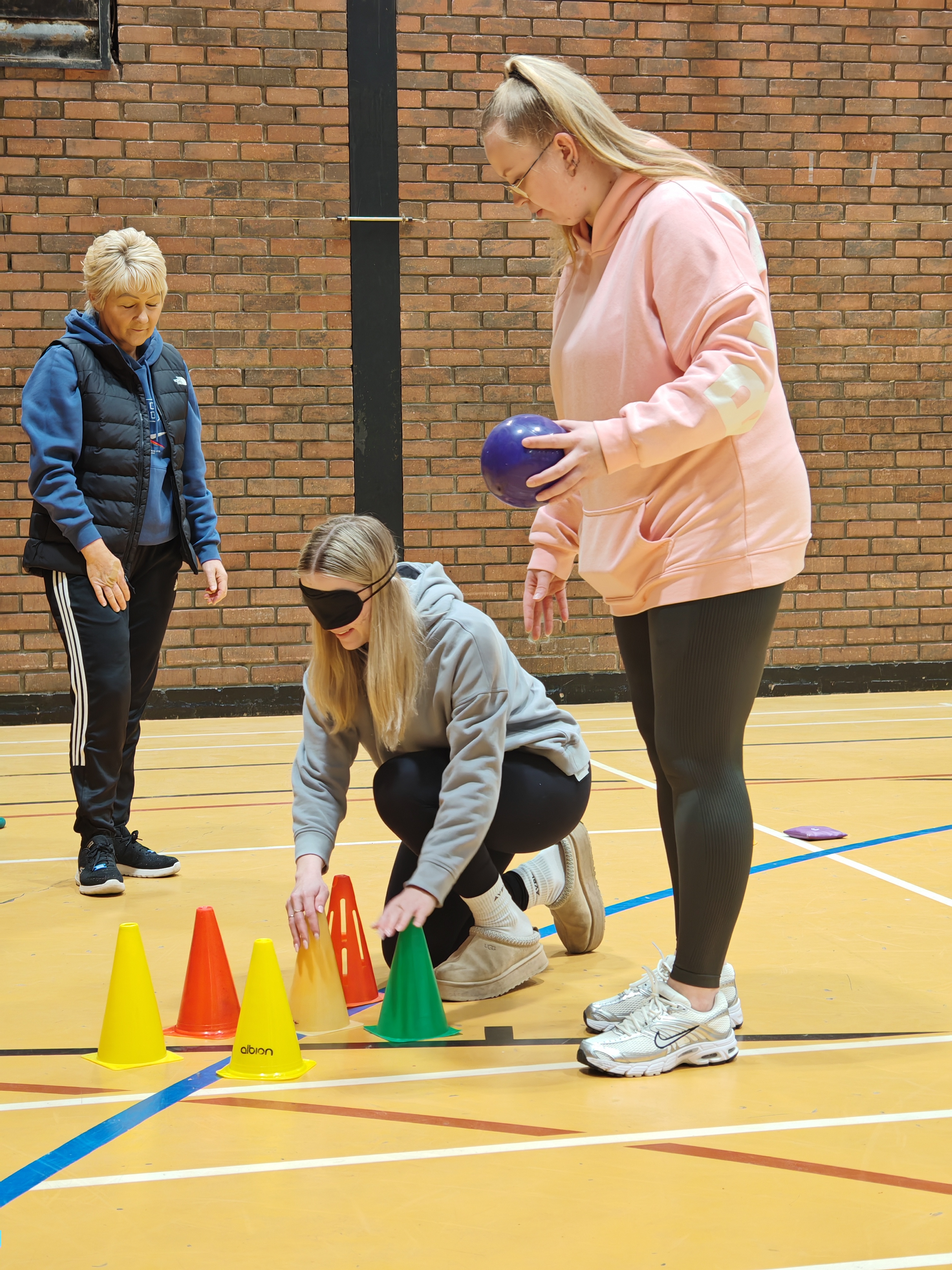 3 people in sports hall. 1 is blind folded,  bending down to feel some cones in front of her. Another is stood next to her watching, holding a ball. The third is stood back watching