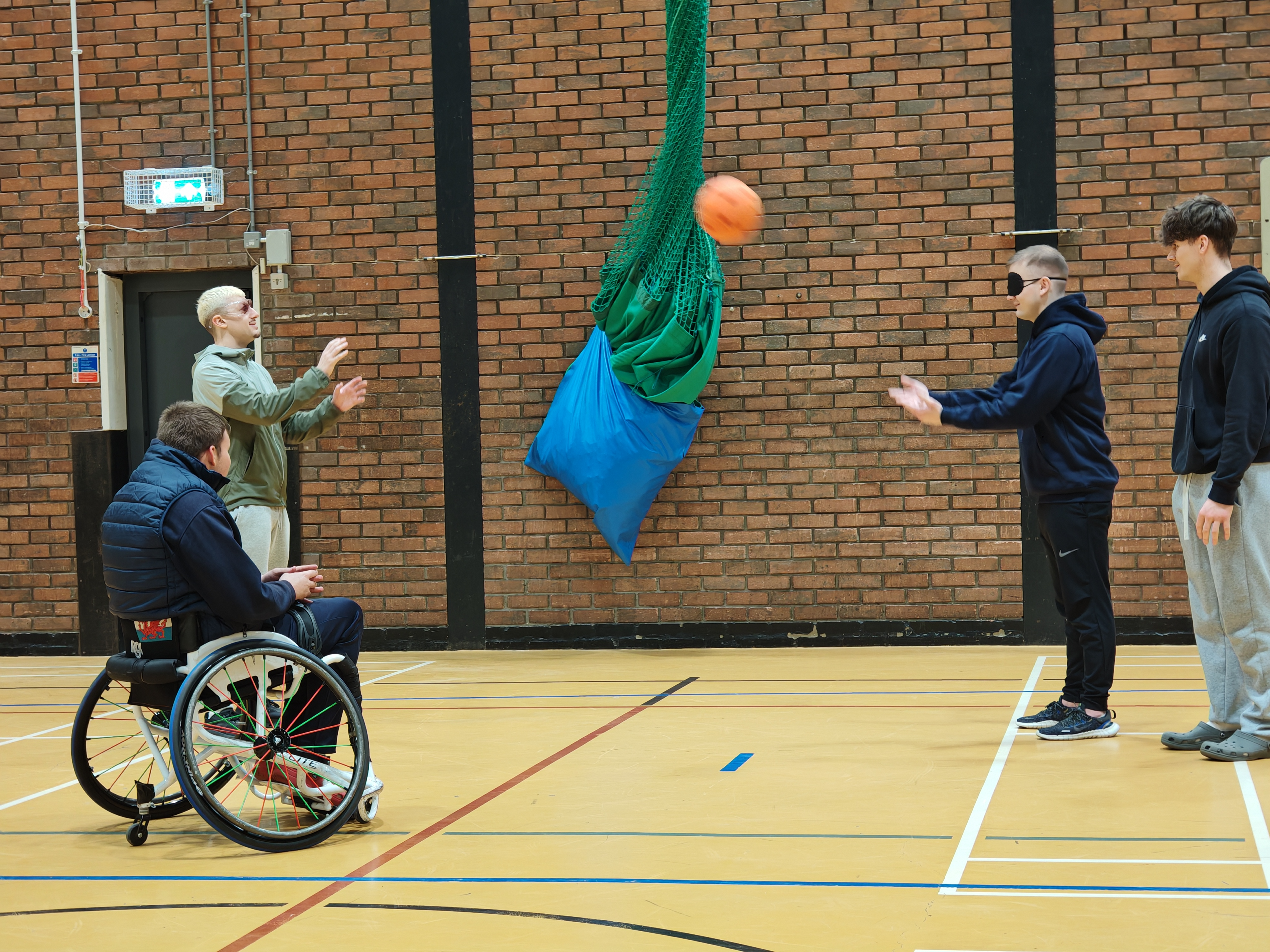 4 Men playing catch in a sports hall. One is in a wheelchair and one is blindfolded