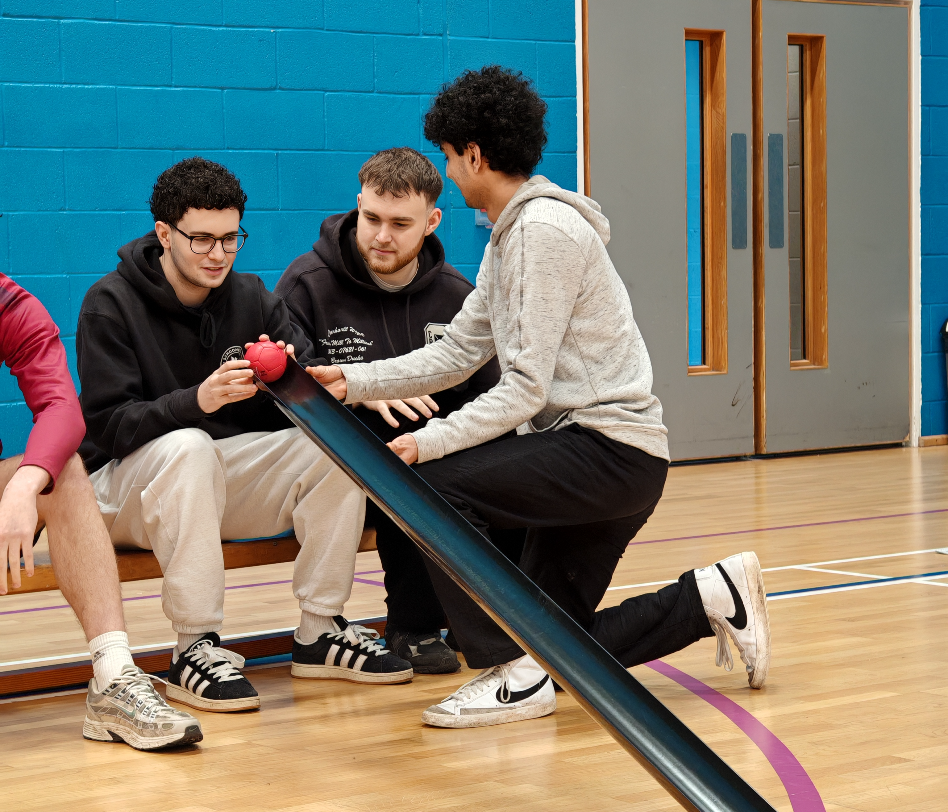 3 males sitting on a bench in a sports hall playing boccia. 1 person is crouch in front holding a black ramp. 1 person is putting the red ball on the ramp and looking forwards to aim the ball