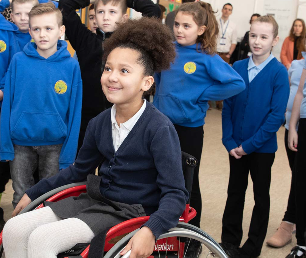 A group of school children aged 10 - 11 in a sports hall, looking towards a teacher. 1 girl is in a sports wheelchair